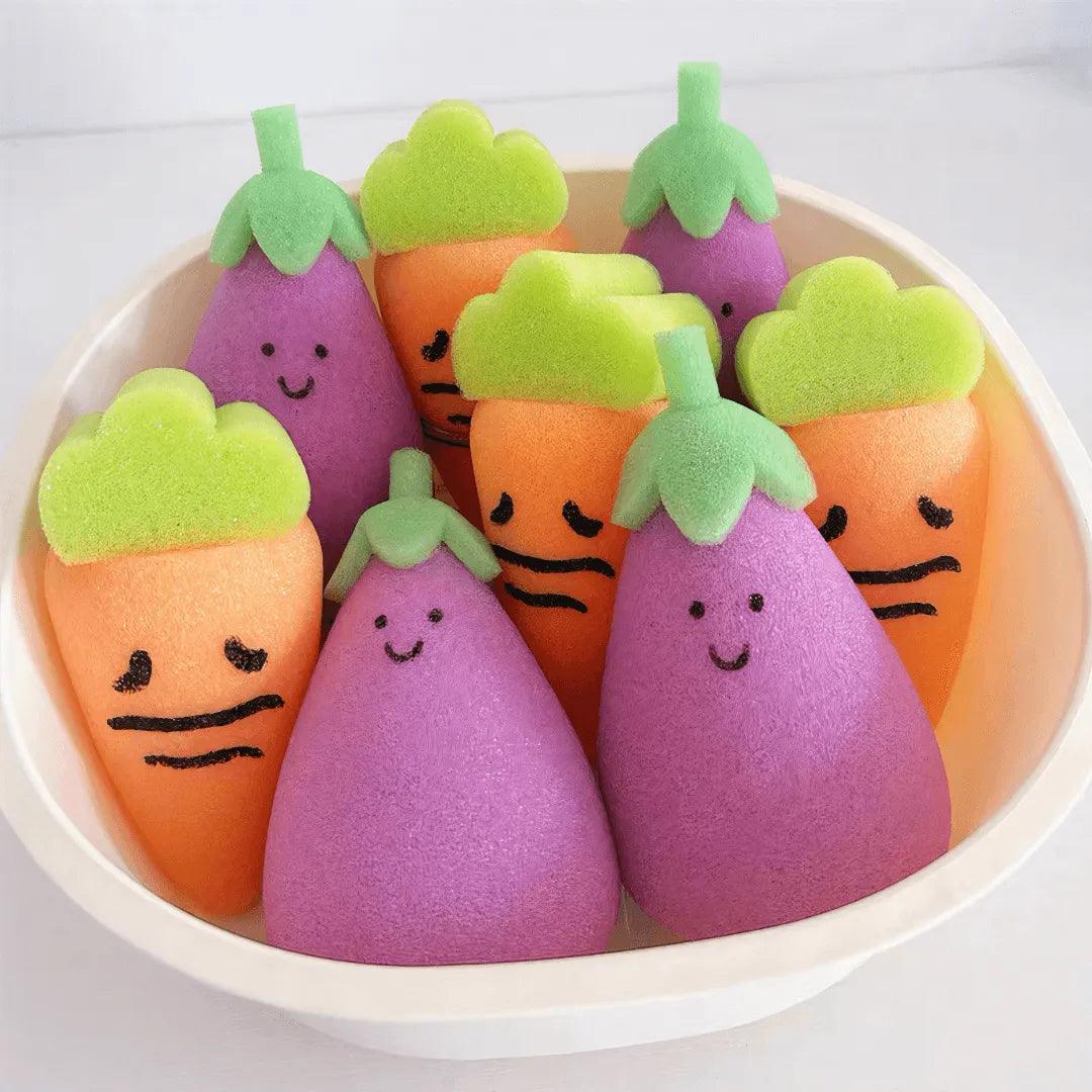 Colorful carrot and eggplant-shaped sponges with cute faces arranged in a white basin.