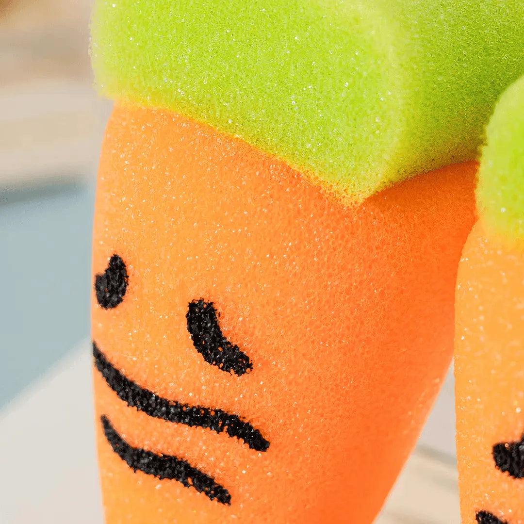 Close-up of orange carrot-shaped sponge with green top showing soft PU texture and cute face.