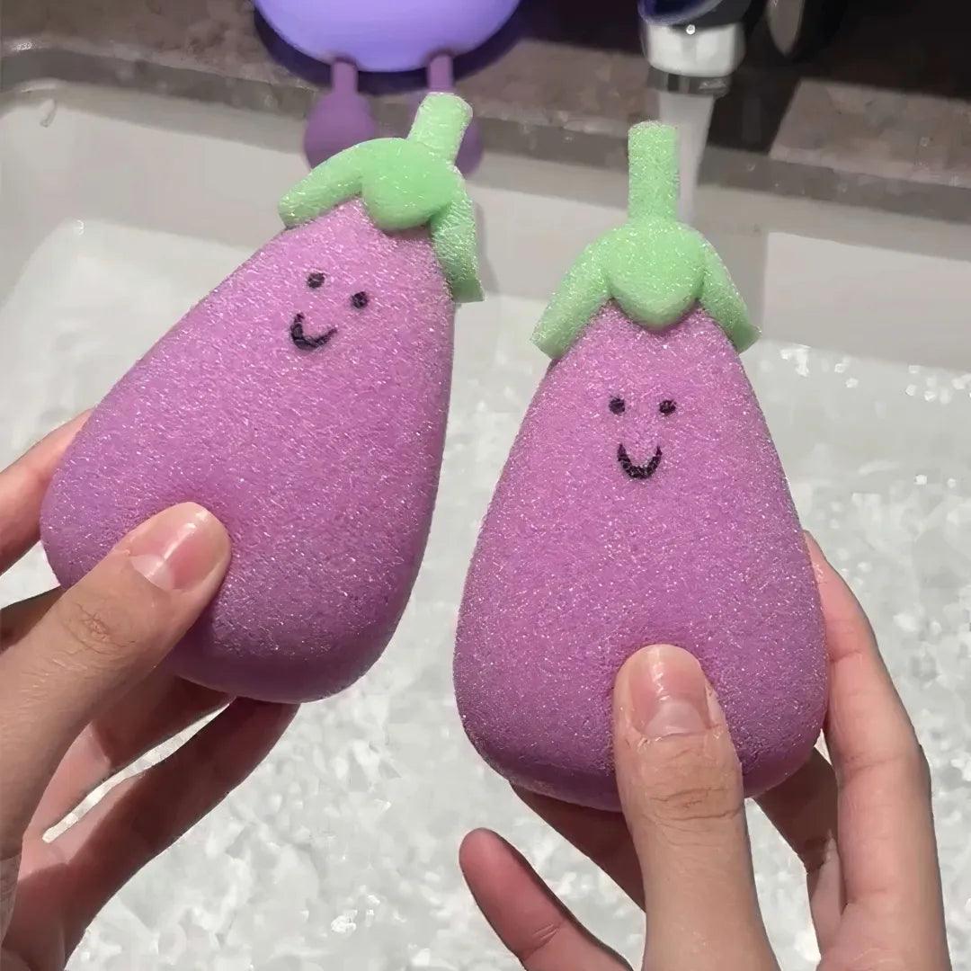 Two purple eggplant sponges with green tops held in hands near sink, textured and smiling.