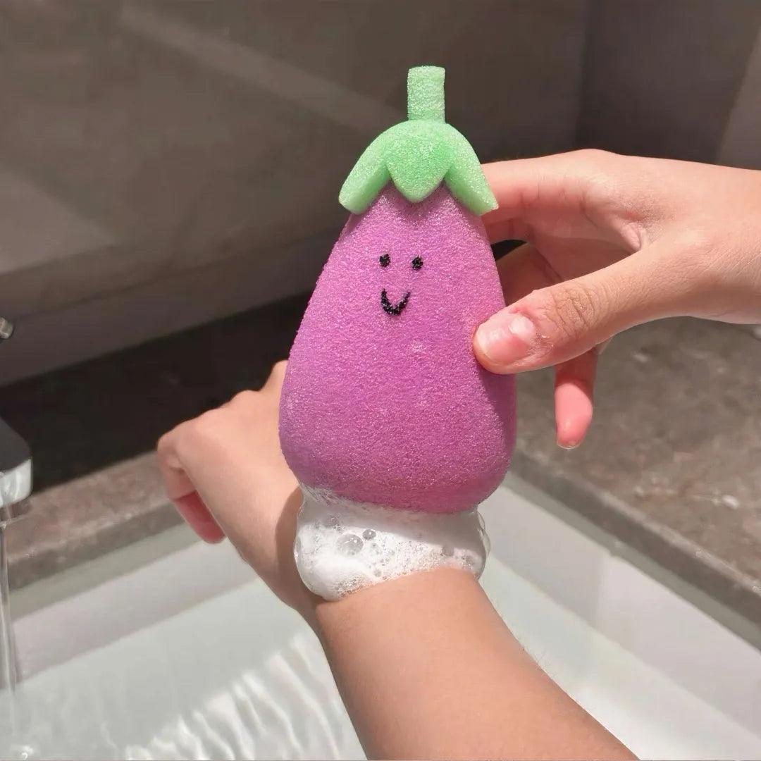Purple eggplant-shaped sponge with green top being used on wrist, foamy and soft texture.
