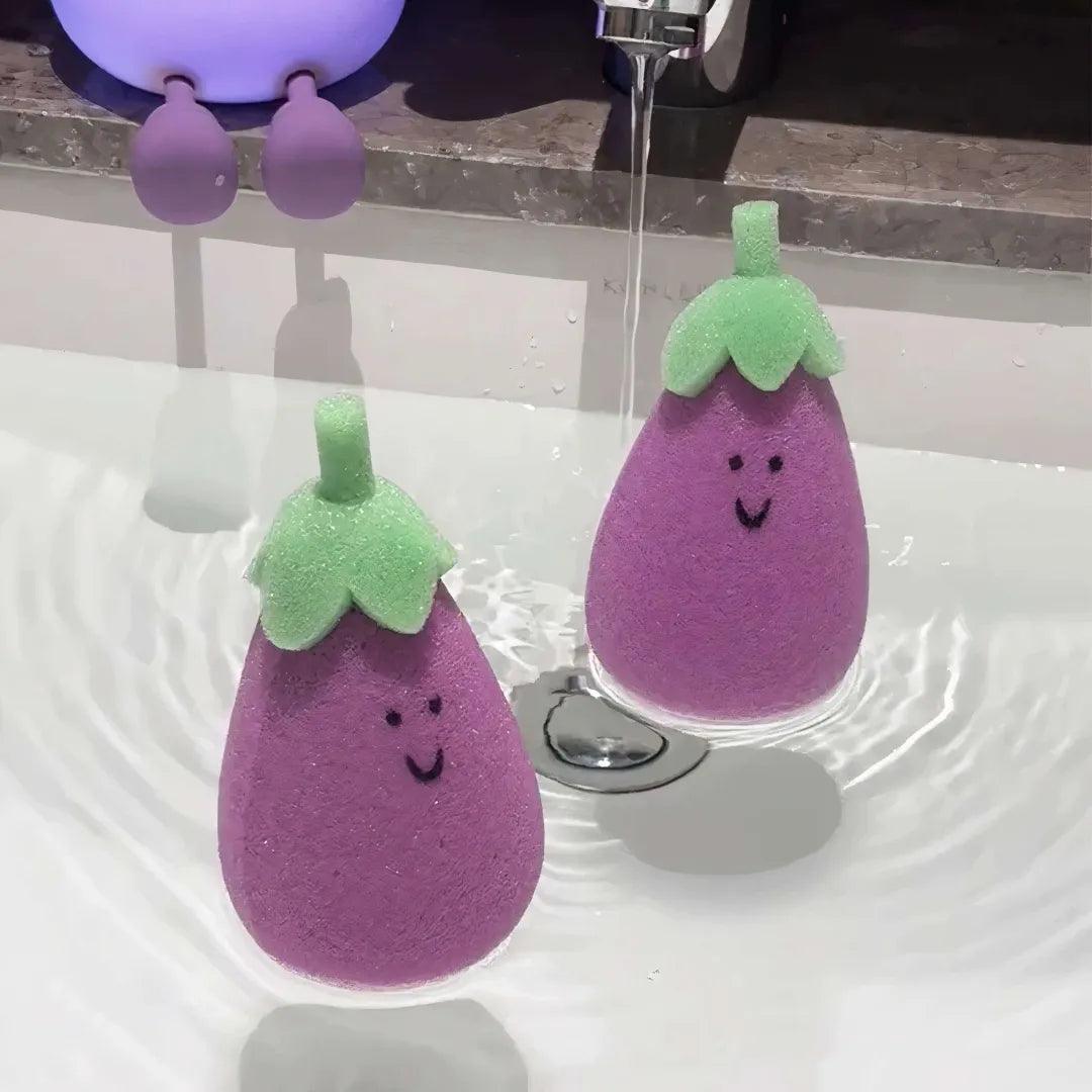 Two purple eggplant-shaped sponges with green tops floating in water under running tap.