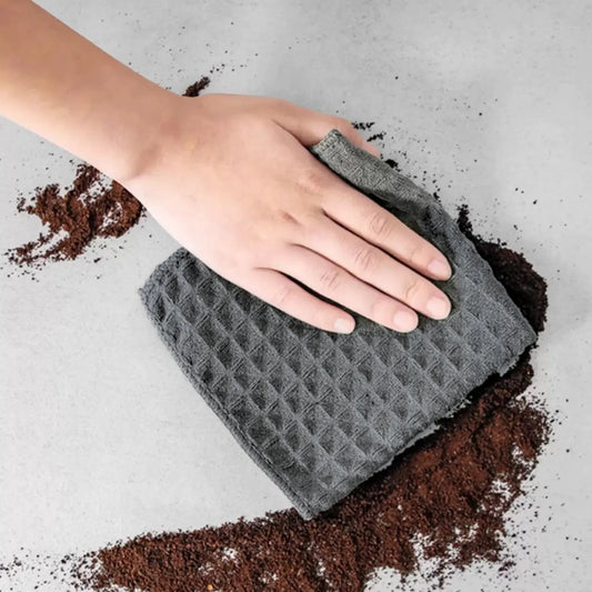 A person wipes coffee grounds off a table using a waffle pattern microfiber cloth; the cleaned area is spotless, showcasing its super absorbent and streak-free performance.
