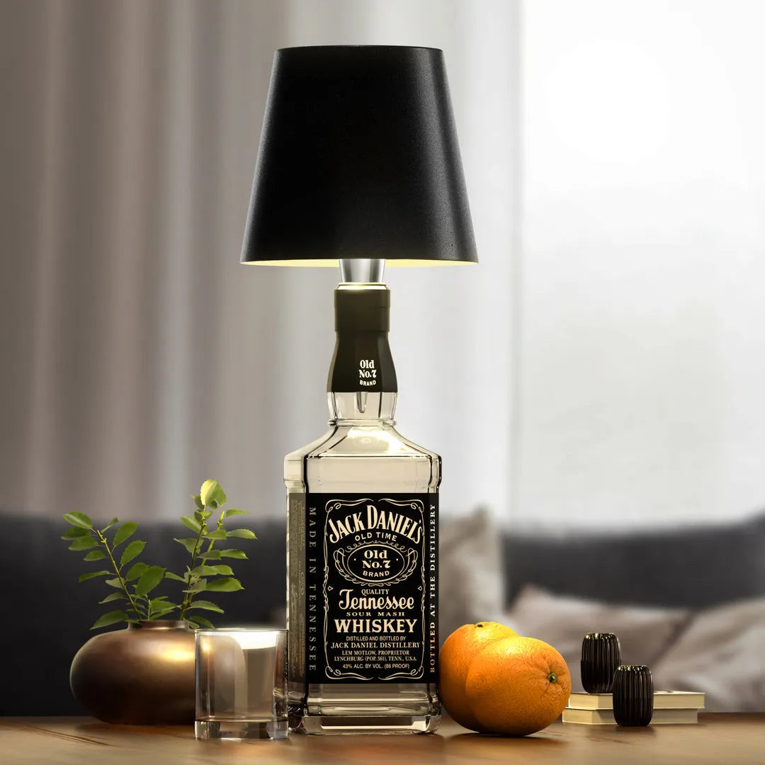 A rustic and creative Jack Daniels whiskey bottle table lamp with a black lampshade, placed on a wooden table beside a glass, oranges, and potted plants.