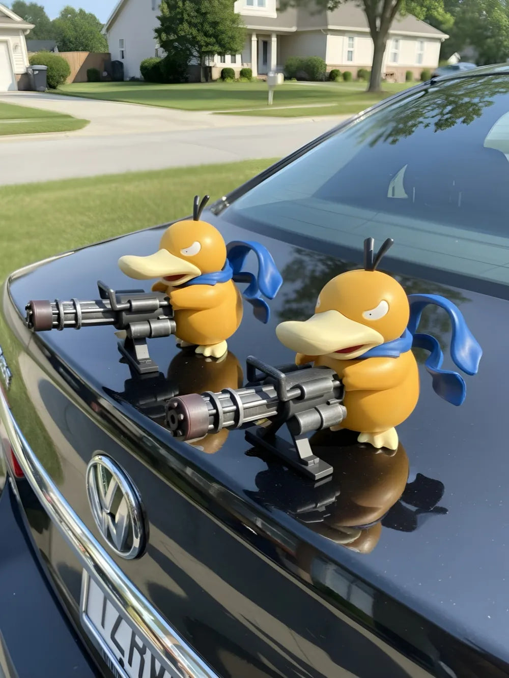 Two Psyduck car ornaments with machine guns mounted on a black Volkswagen hood, set against a sunny suburban street—a quirky gift for car enthusiasts.