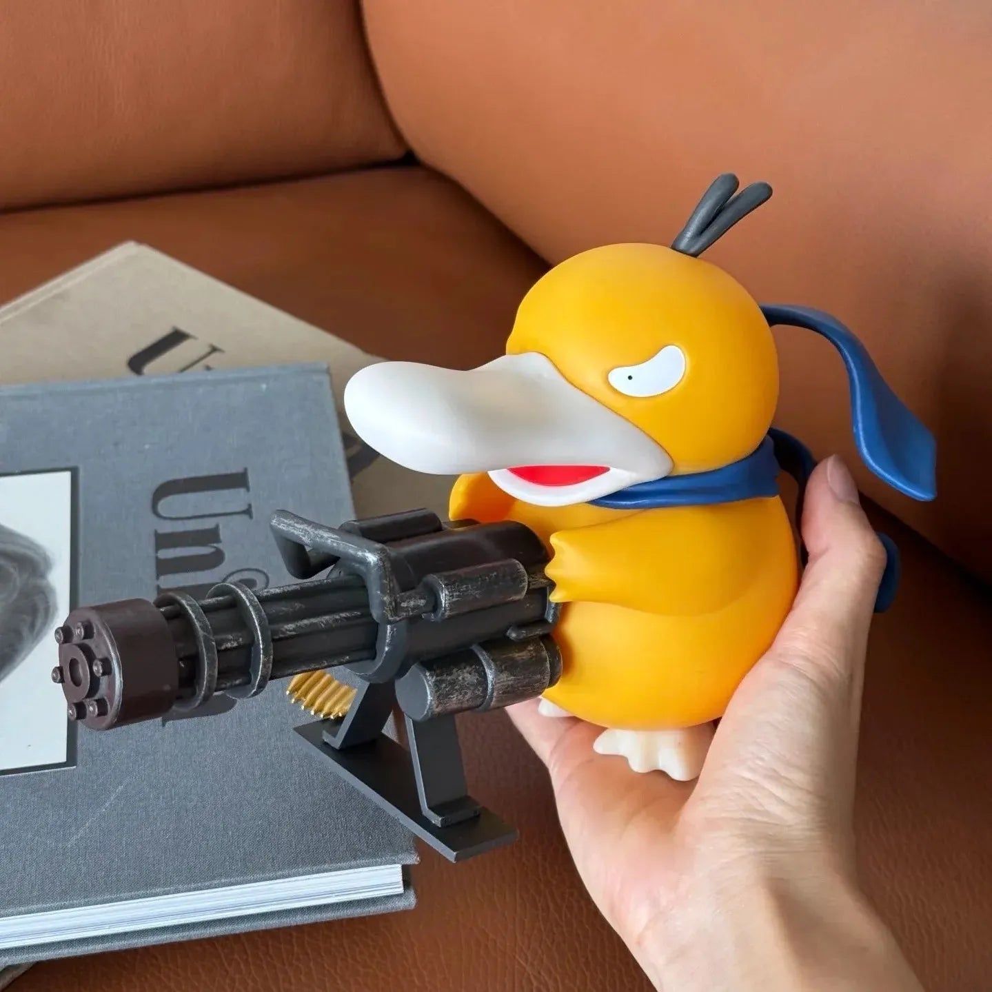 Hand holding a small yellow duck-like figurine with a blue bow tie and black weapon on a brown surface.