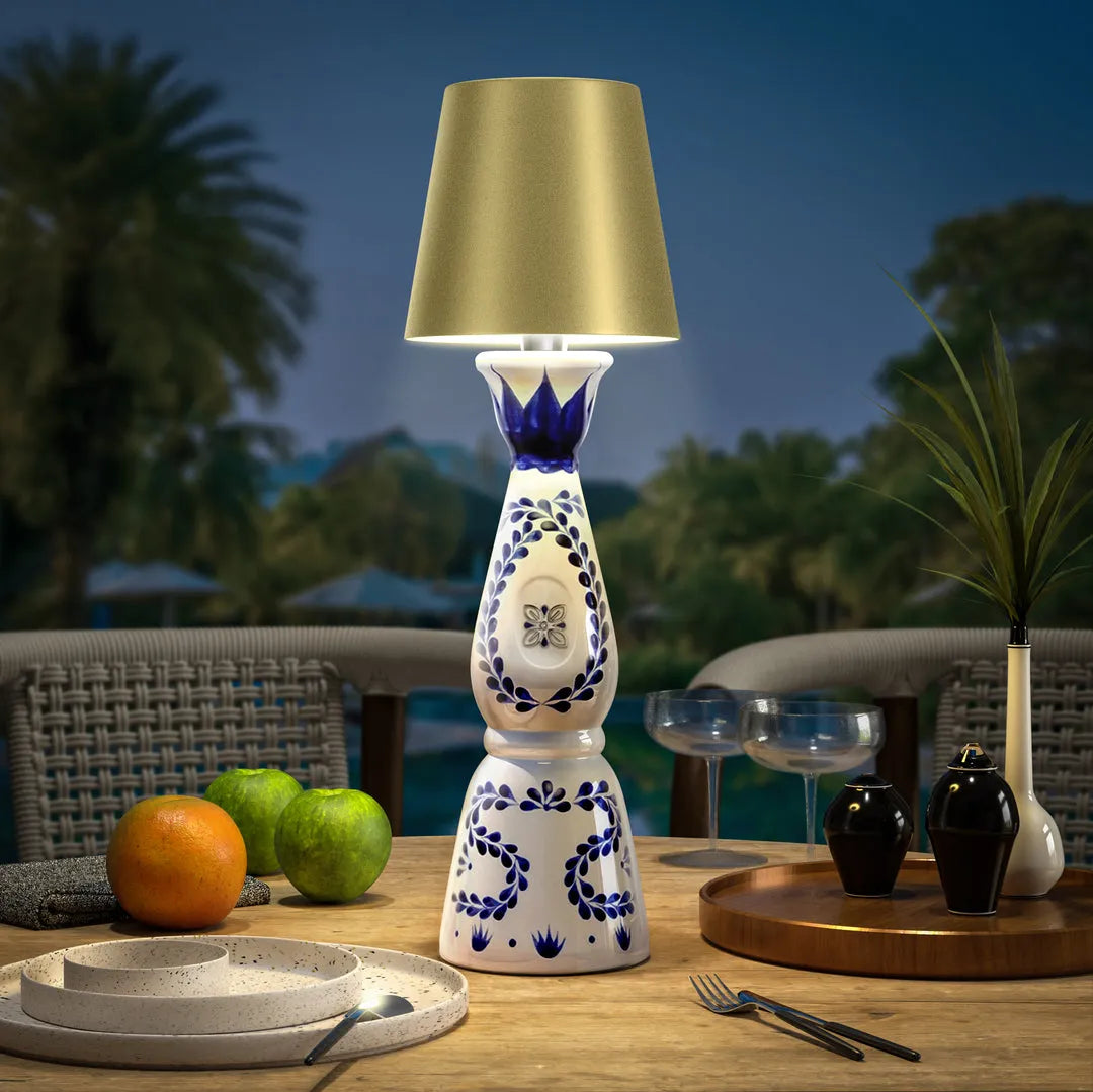Wireless bottle lamp with intricate blue and white patterns and a golden lampshade, placed on a wooden outdoor table beside fresh fruits and glassware.