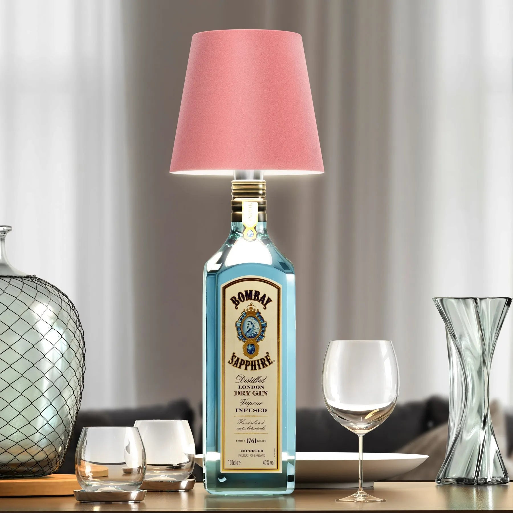 Wireless bottle lamp made from a Bombay Sapphire gin bottle with elegant pink lampshade, placed on a table with wine glasses and vases.