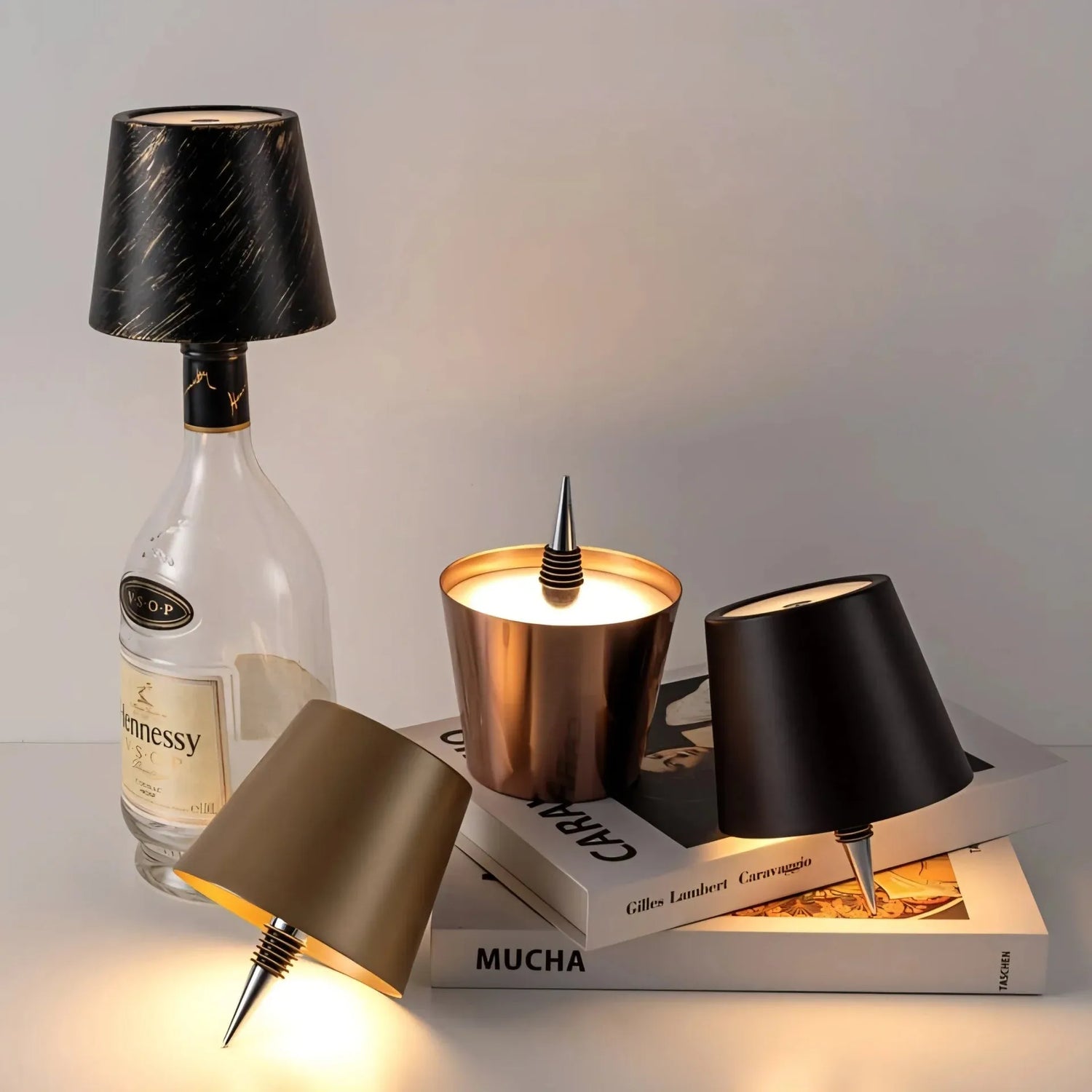 Wireless Hennessy bottle lamp with black and gold streaked shade, paired with bronze and matte black mini lamps on stacked books.
