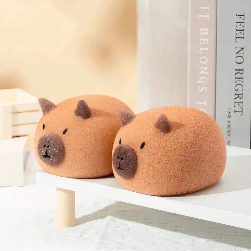 Two round light brown capybara-shaped sponges with dark snouts and ears on a white shelf.