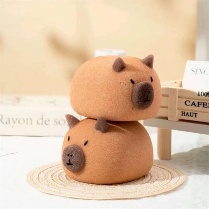 Stacked brown capybara-shaped sponges with dark ears and nose on woven mat and table.