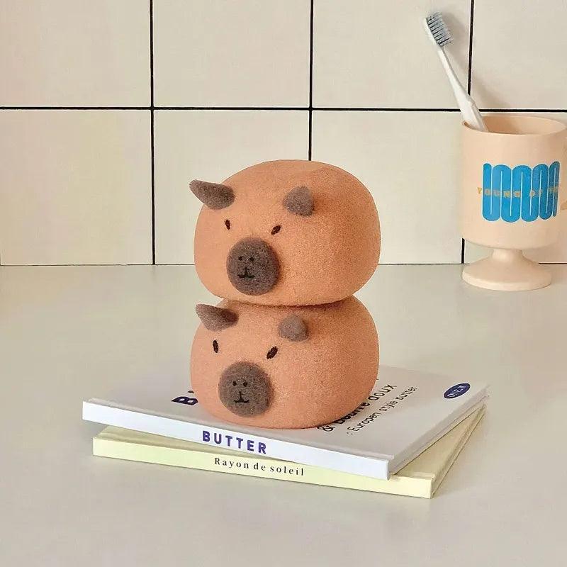 Two light brown capybara-shaped sponges with dark snouts stacked on books in bathroom setting.


