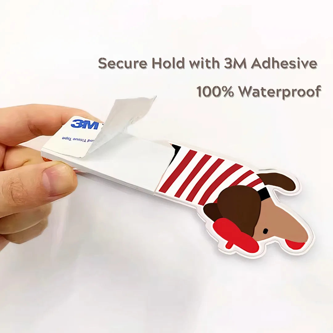 A dachshund-shaped toilet seat lifter with red stripes, featuring strong 3M adhesive backing, being peeled by hand for easy and secure installation.