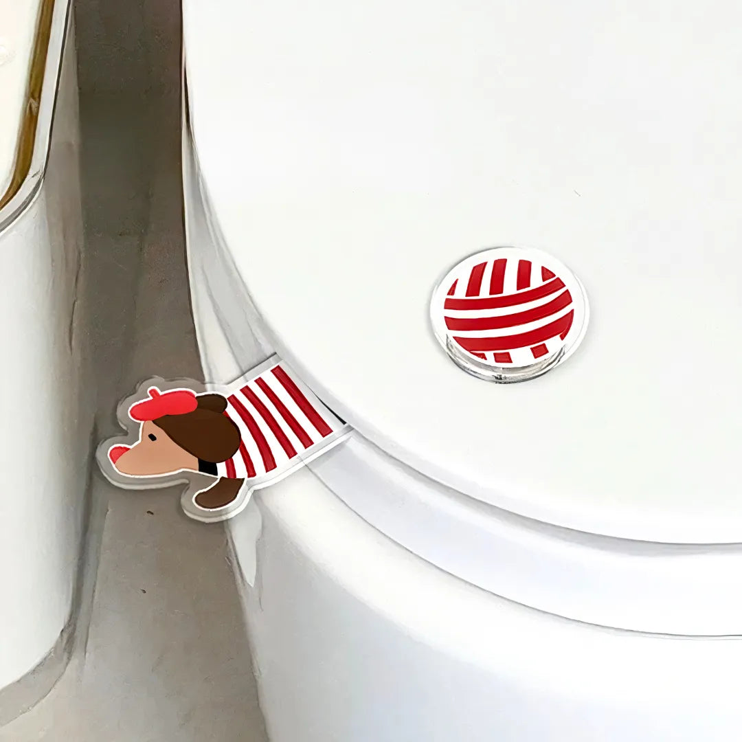 A dachshund-shaped toilet seat lifter made of acrylic, featuring a red striped outfit, attached to the side of a white toilet lid.