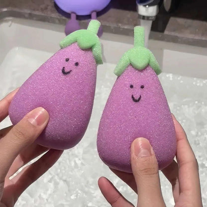 Two purple eggplant sponges with green tops held in hands near sink, textured and smiling.