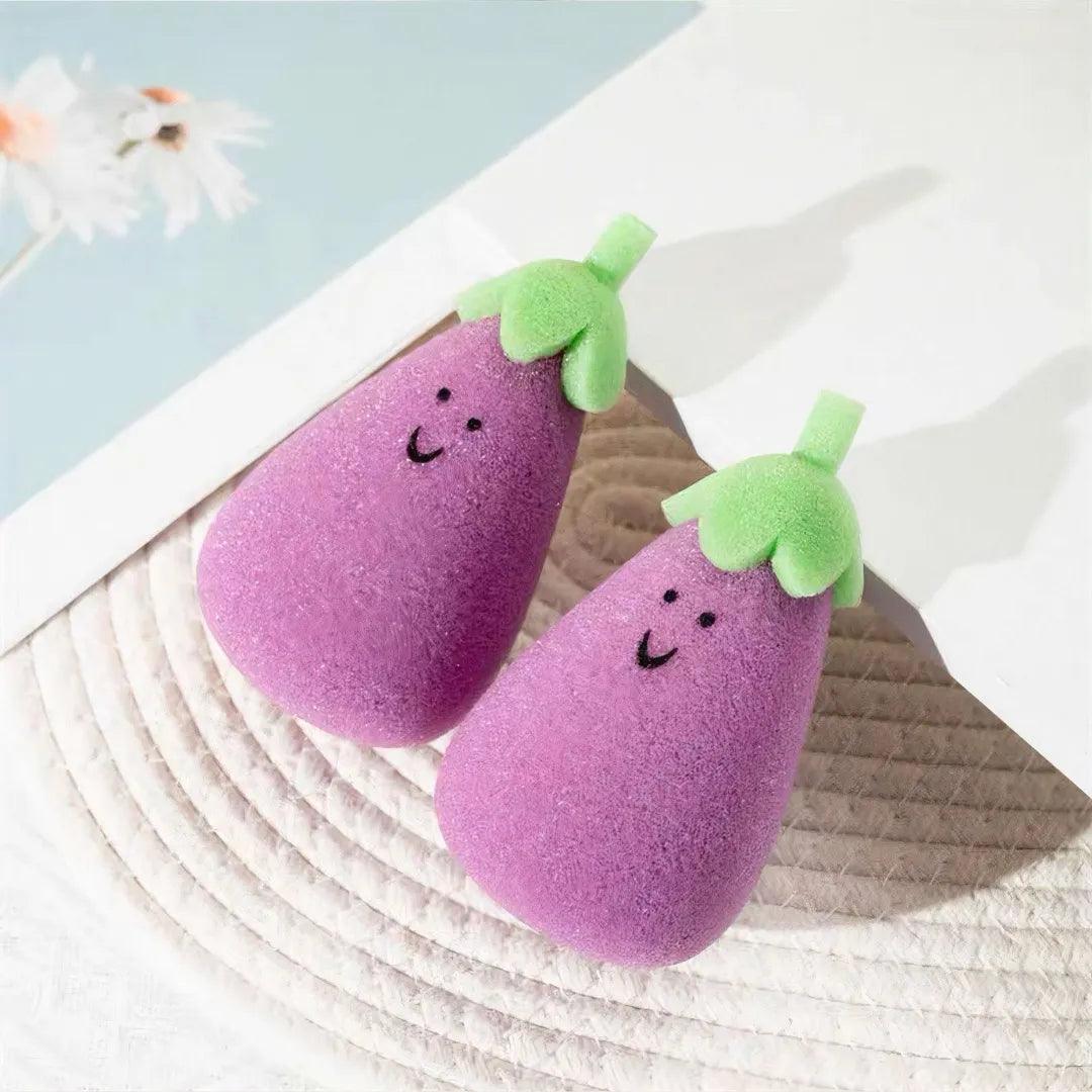 Two purple eggplant-shaped sponges with green tops and smiley faces on light woven mat.