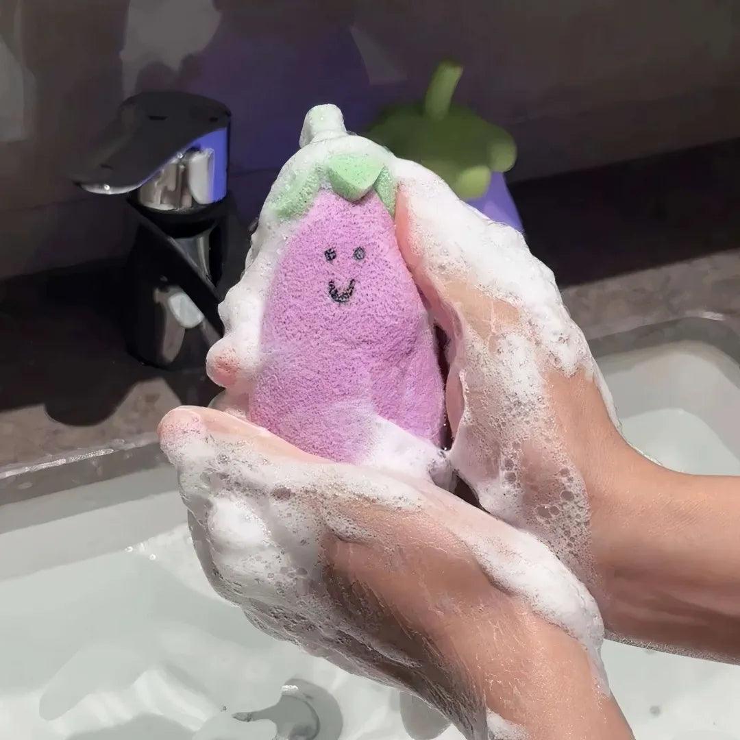 Purple eggplant-shaped sponge with green top covered in rich foam between two hands.