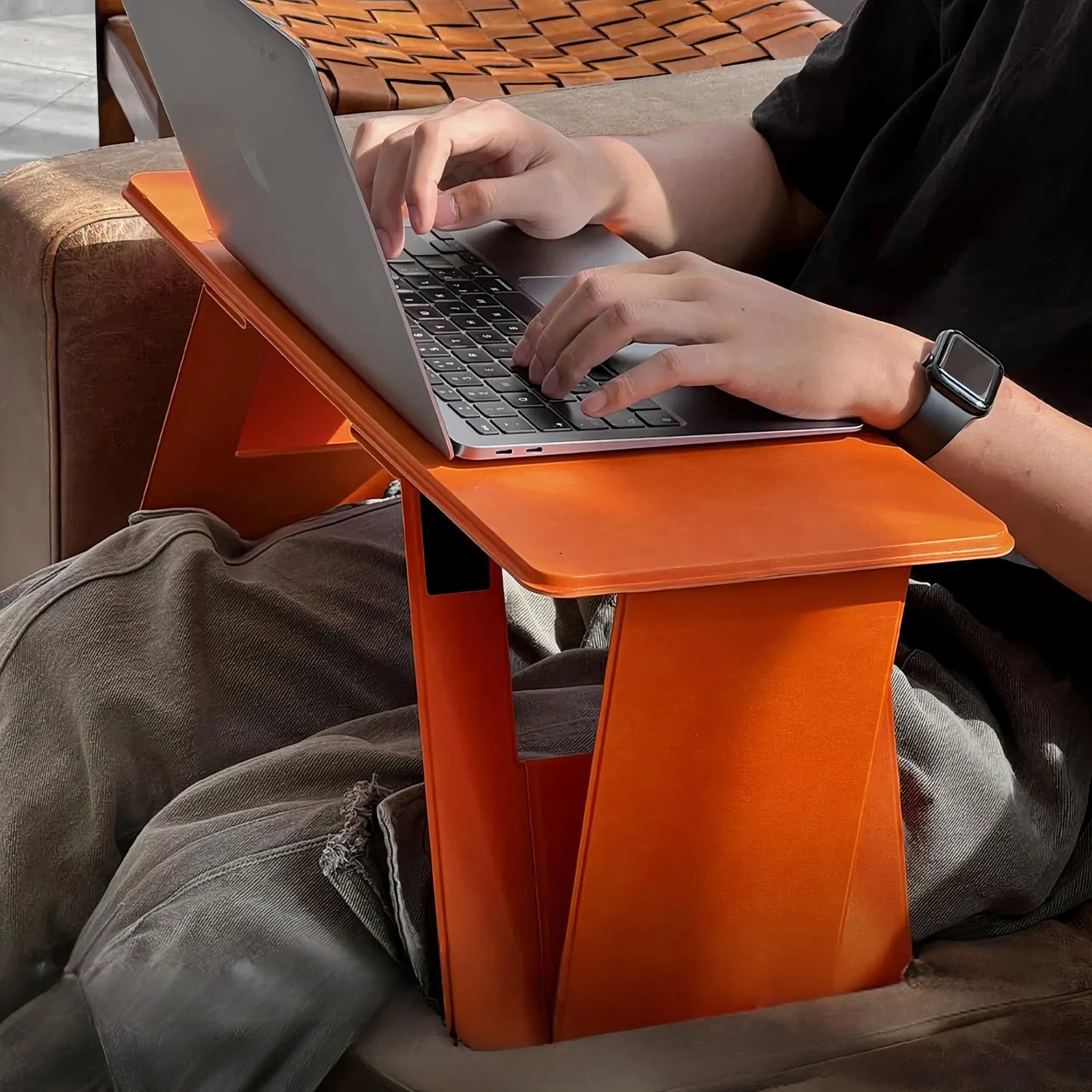 A person is using a slim, foldable lap desk with a laptop while sitting on a sofa. The lightweight yet sturdy structure offers a stable work surface and folds flat for portability.