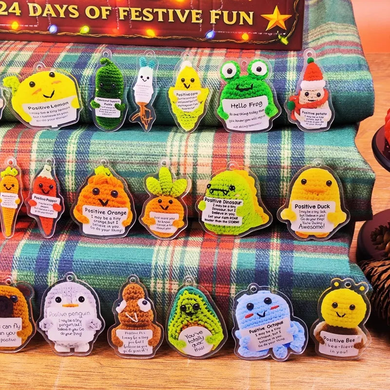 2025 Emotional Support Advent Calendar open to show day 24's amigurumi ornament with smile, placed on red plaid fabric beside the closed box.