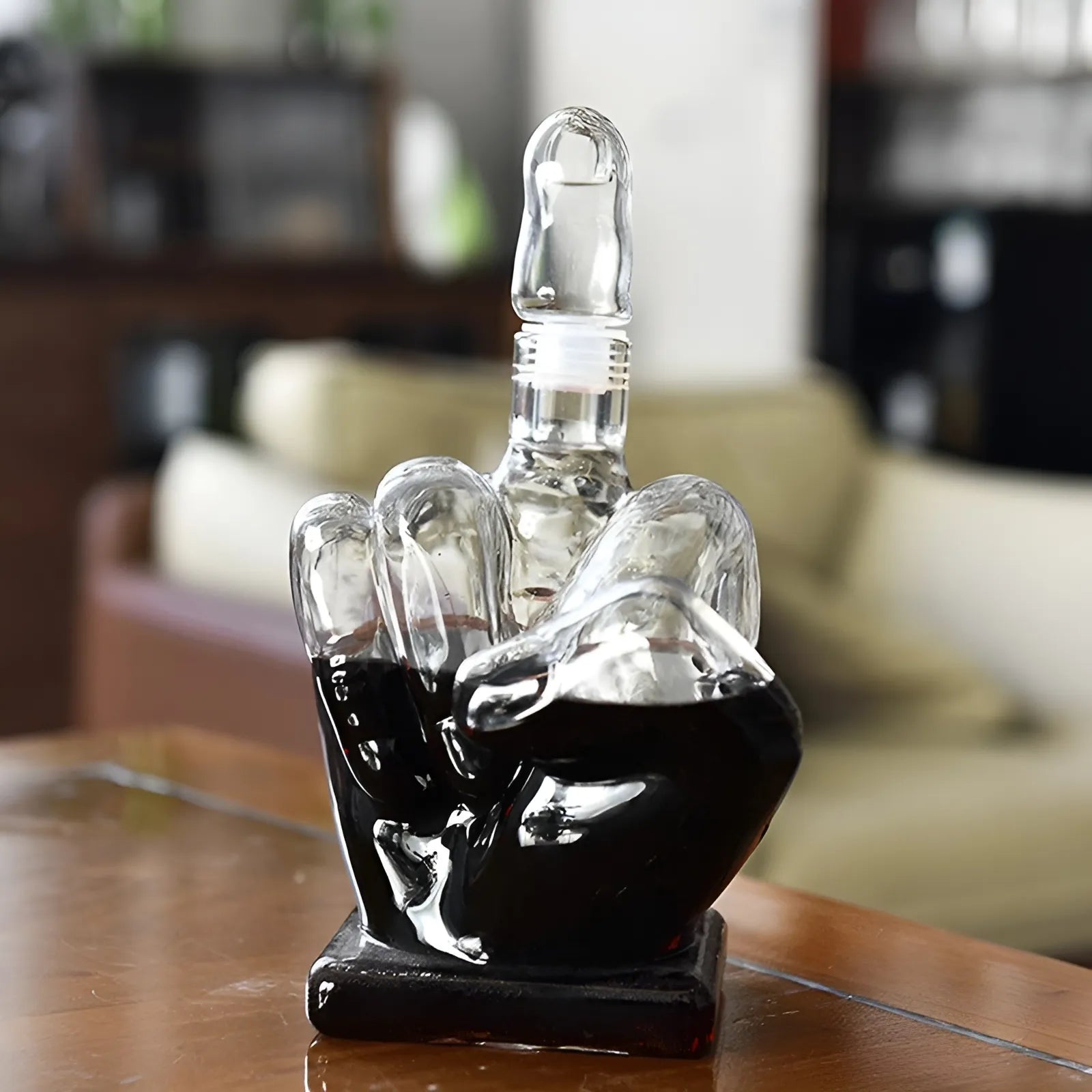 Glass middle finger decanter with wine on wooden table. Blurred living room background highlights its unique shape.