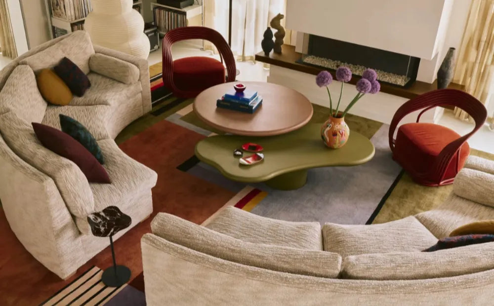 Modern living room featuring a curved beige sofa with colorful cushions, organic-shaped coffee tables, red accent chairs, and a fireplace with decorative mantel. Stylish contemporary interior design.