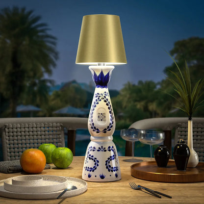 Wireless bottle lamp with intricate blue and white patterns and a golden lampshade, placed on a wooden outdoor table beside fresh fruits and glassware.