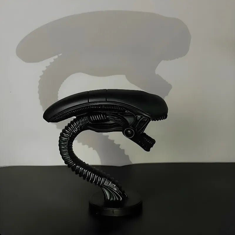 Xenomorph alien desk lamp casting dramatic shadow on white wall. Shows detailed biomechanical design with curved silhouette and circular base.